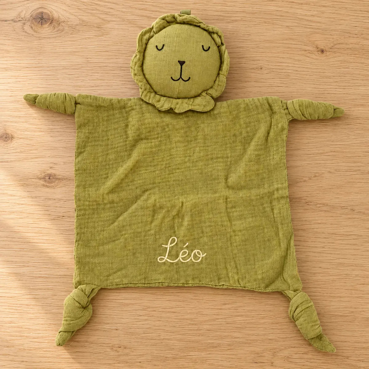 Doudou lion personnalisé vert olive présenté à plat sur du bois clair dans une ambiance nature