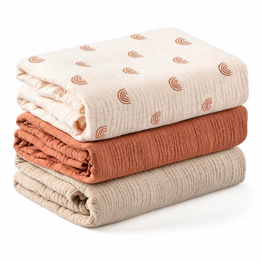 Lot de trois couvertures bébé pliées : écru à motifs arc-en-ciel, terracotta uni et beige uni.