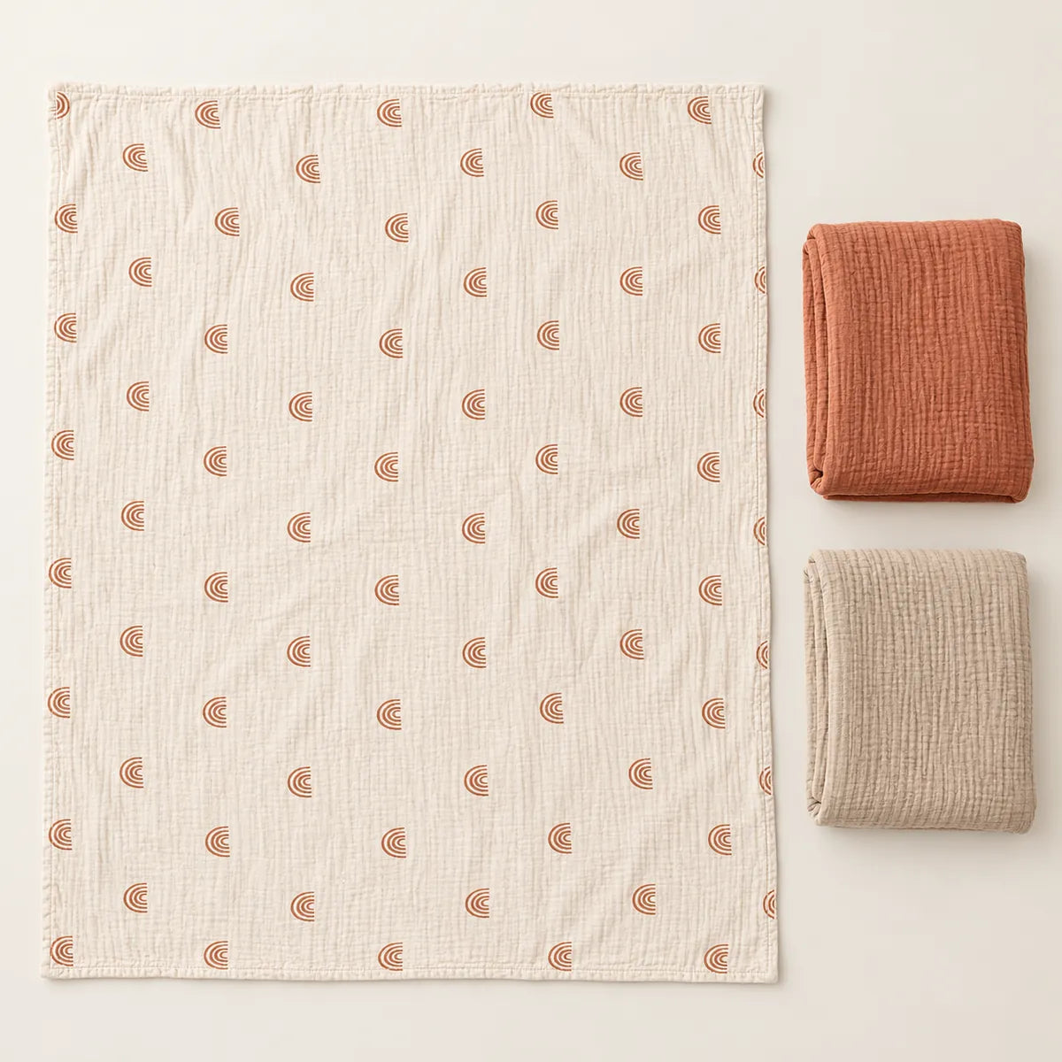 Couverture écrue à arcs-en-ciel dépliée à côté des couvertures terracotta et beige pliées.
